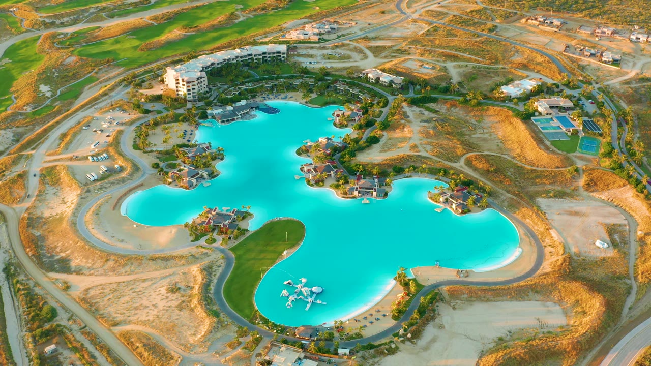 Aerial frame of the Diamante Cabo San Lucas man-made lake with bright blue water centered, small islands, surrounding access roads, dry construction land, buildings, and no visible people.