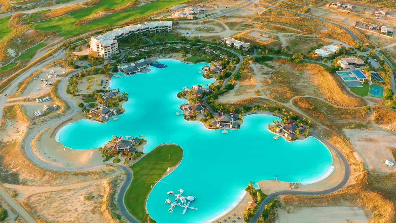 Opening aerial frame over a turquoise lagoon at Diamante Cabo San Lucas, with sandy roads, resort buildings, green landscaping, and no visible people.