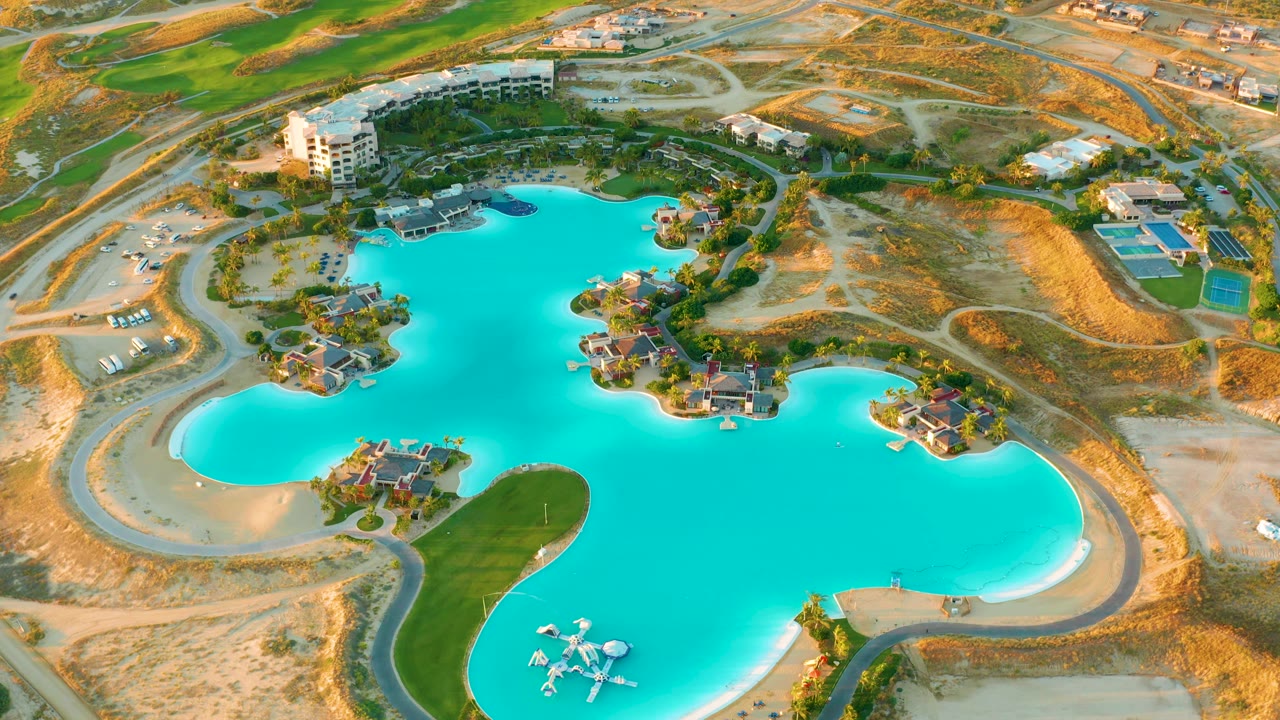 At 0:02, the aerial view shows bright lagoon water, clustered resort structures along the shore, pale desert terrain, and no visible people in Diamante Cabo San Lucas.