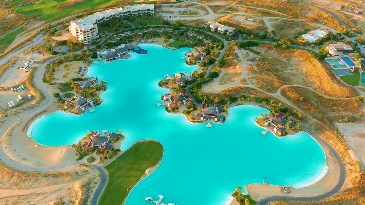 Second aerial frame in Diamante Cabo San Lucas with the blue lake dominating the lower view, houses on landscaped islands, roads curving through dry terrain, and no visible people.