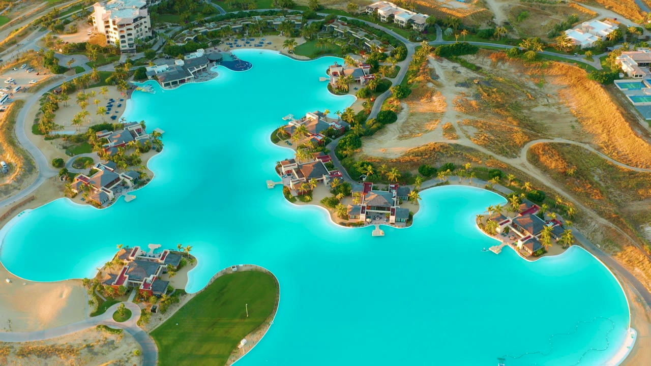 Aerial frame over Diamante Cabo San Lucas showing bright blue lake water, small house islands, sandy edges, roads, dry ground, and no visible people.