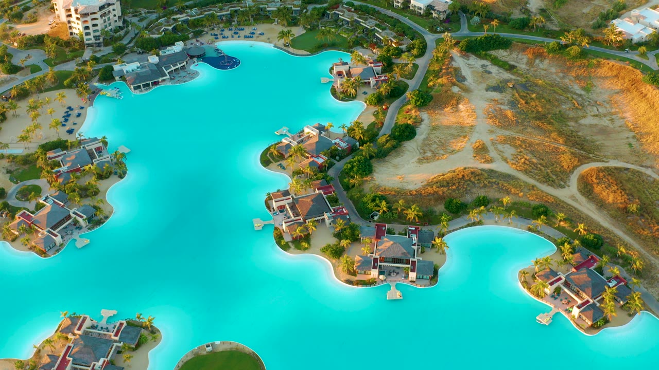 Opening overhead frame of a turquoise lagoon at Diamante Cabo San Lucas, Mexico, bordered by villas, palms, roads, and dry sandy terrain, with no visible people.