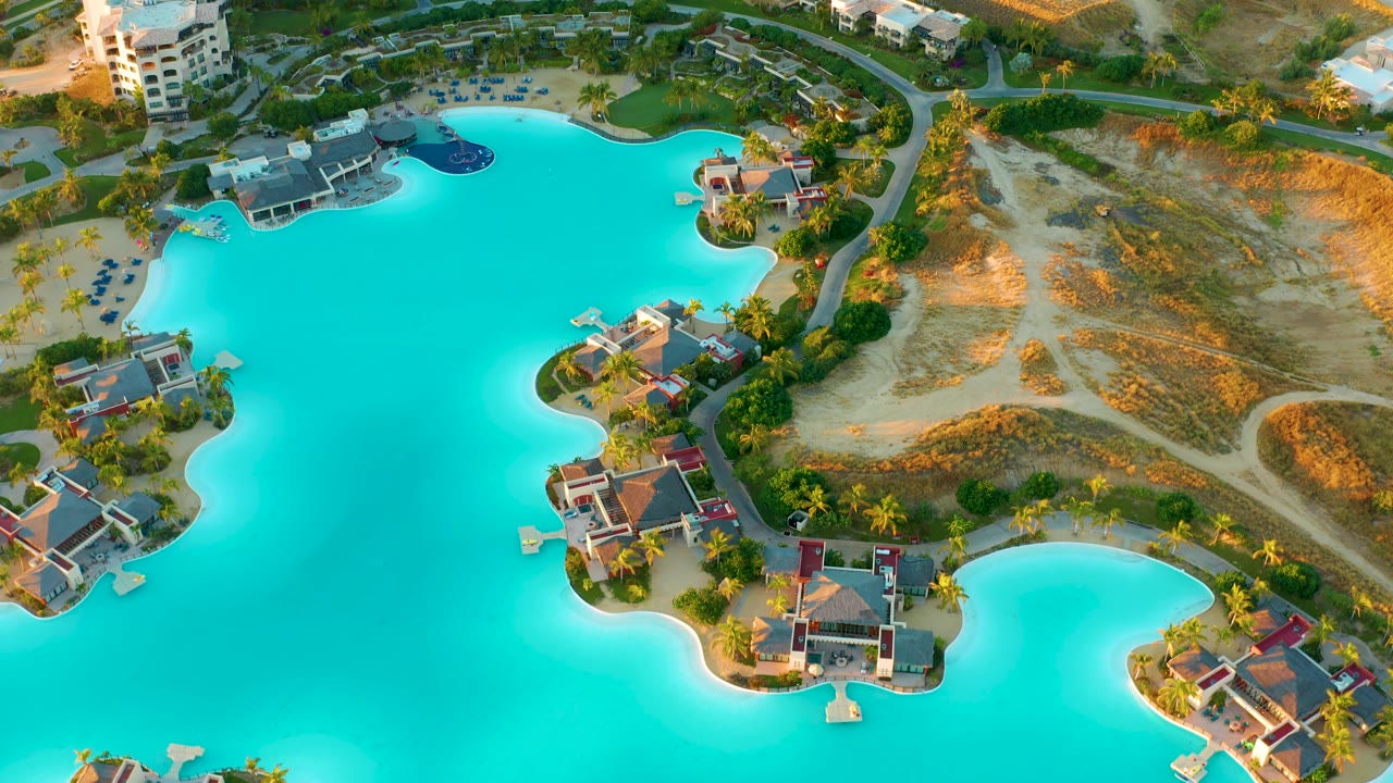 Aerial lagoon scene with resort buildings, sandy ground, palms, and bright blue water.