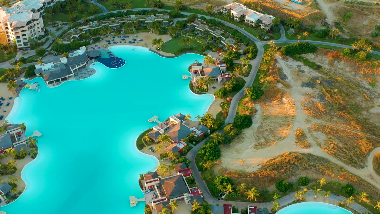 Opening aerial frame over a turquoise resort lagoon at Diamante Cabo San Lucas, with buildings, palms, a curving road, sandy ground, and no visible people.