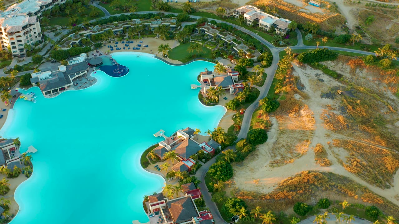 Wide aerial frame over Diamante Cabo San Lucas with turquoise lagoon water, poolside resort buildings, palms, a road, open sandy ground, and no visible people.