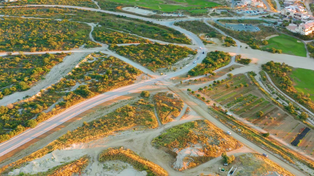 Aerial frame over Diamante Cabo San Lucas showing a pale dirt-road intersection, scrub-covered lots, green fields, and small vehicles, with no visible people.