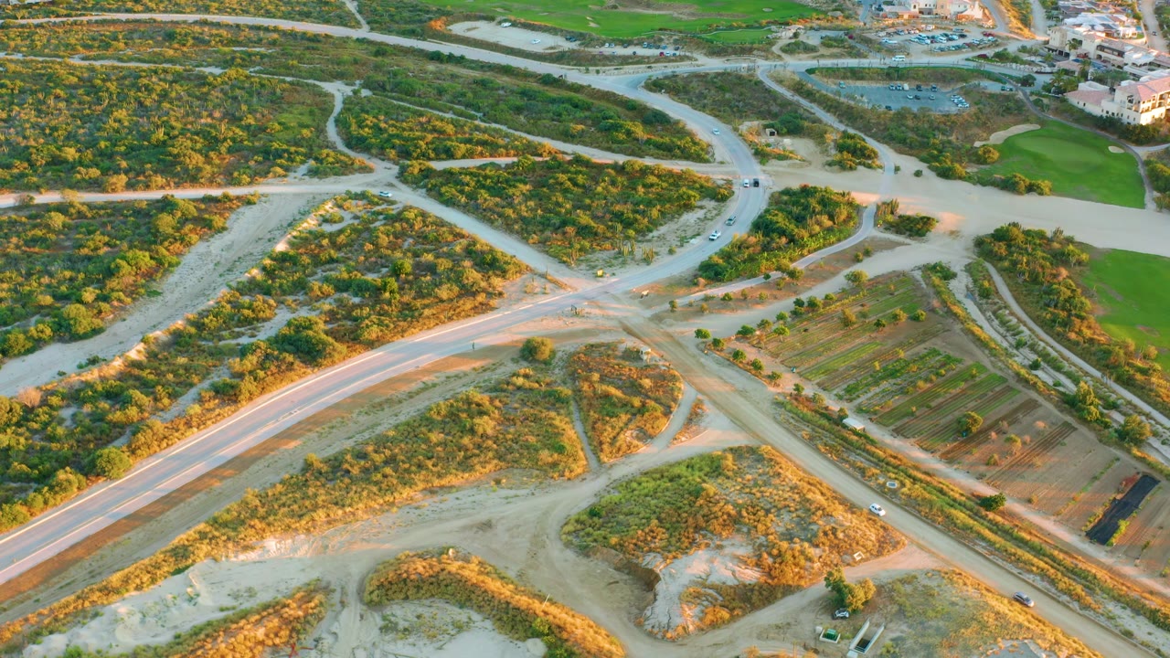 High aerial over Diamante Cabo San Lucas showing a road junction bordered by brush, rectangular graded fields, green areas, and buildings near the top of frame, with no visible people.