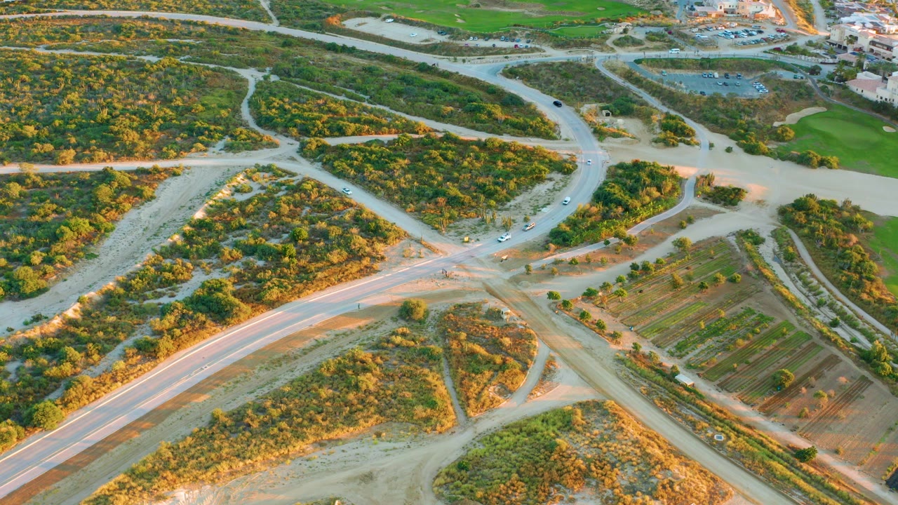 High aerial frame over a light-colored road junction in Diamante Cabo San Lucas, with scrubby green terrain, field rows, a few cars on the roads, and no visible people.
