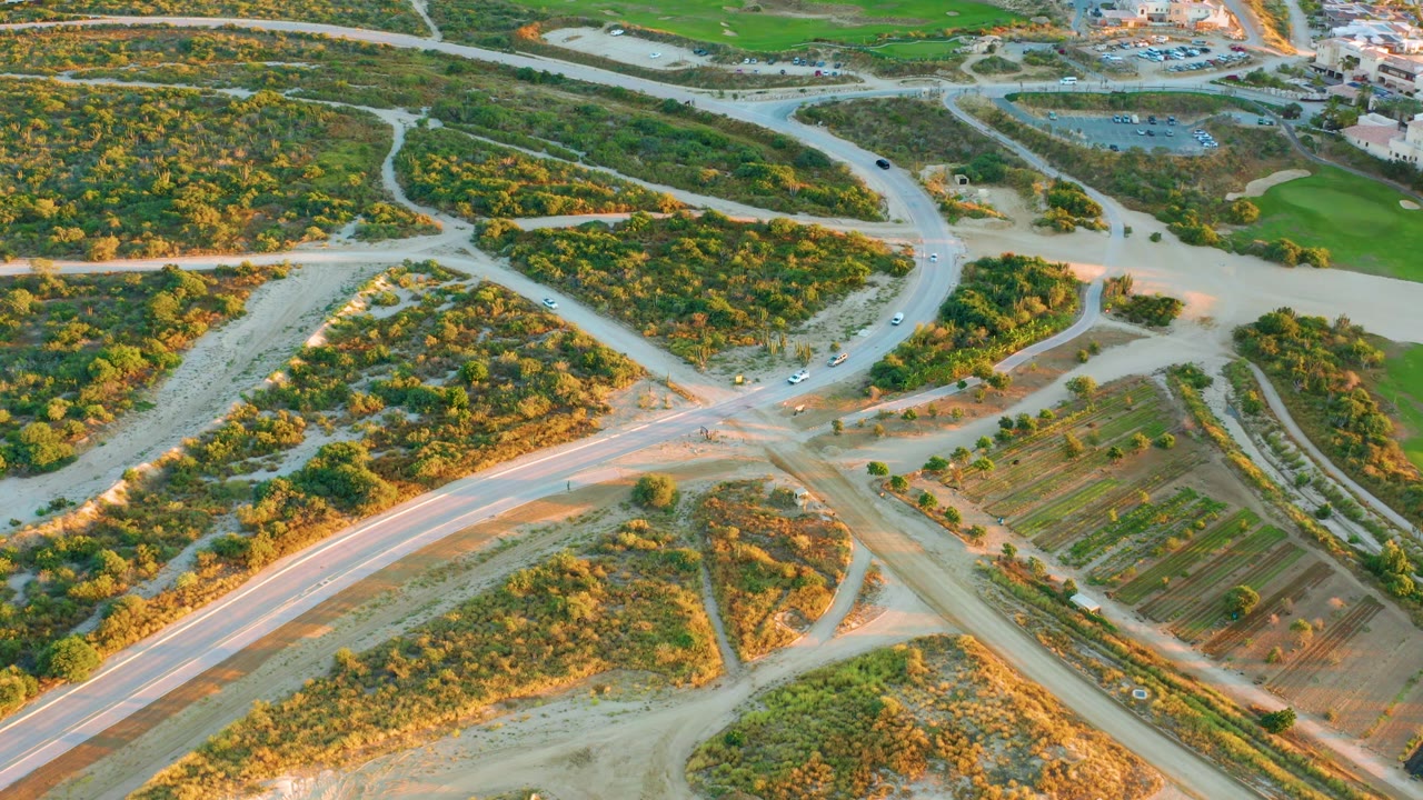 Overhead view of vehicles entering and leaving a pale road intersection in Diamante Cabo San Lucas, bordered by trees, dirt tracks, and planted field rows, with no visible people.