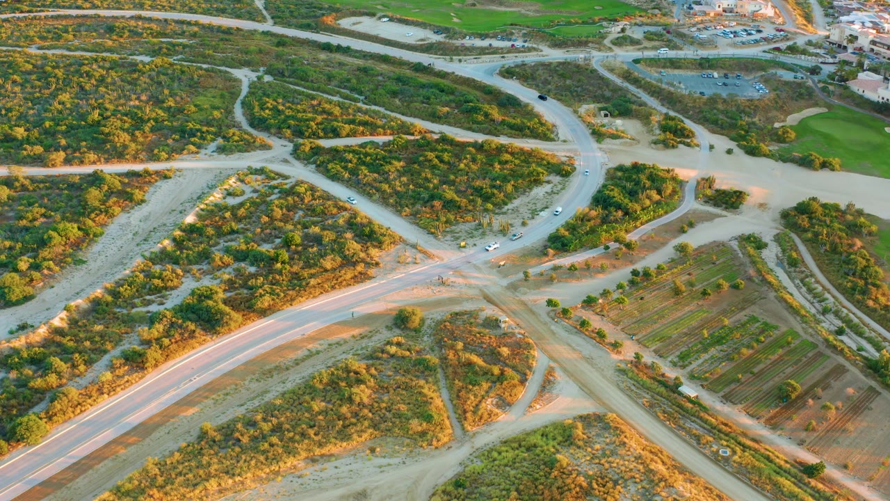 Aerial view in Diamante Cabo San Lucas showing intersecting roads, small vehicles near the junction, green scrub areas, rectangular field rows, and no visible people.