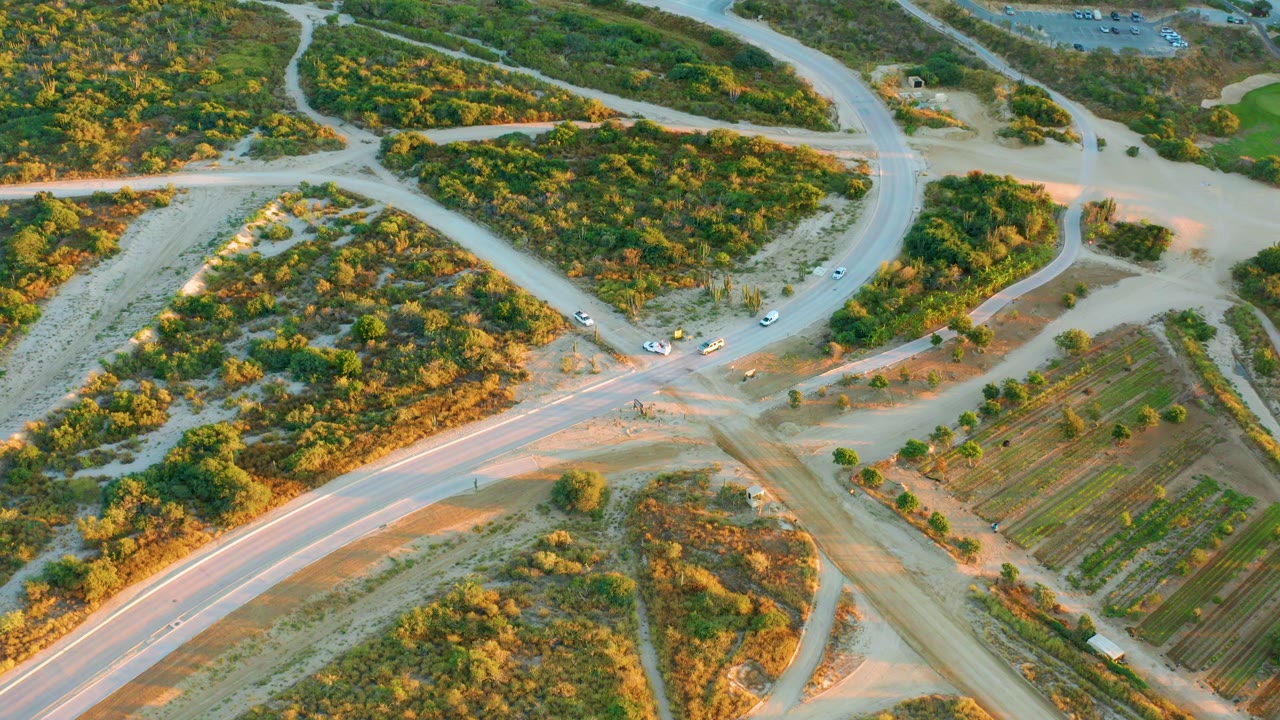 Aerial frame over Diamante Cabo San Lucas showing pale roads meeting among green scrub, cultivated rows at right, several small vehicles near the junction, and no visible people.