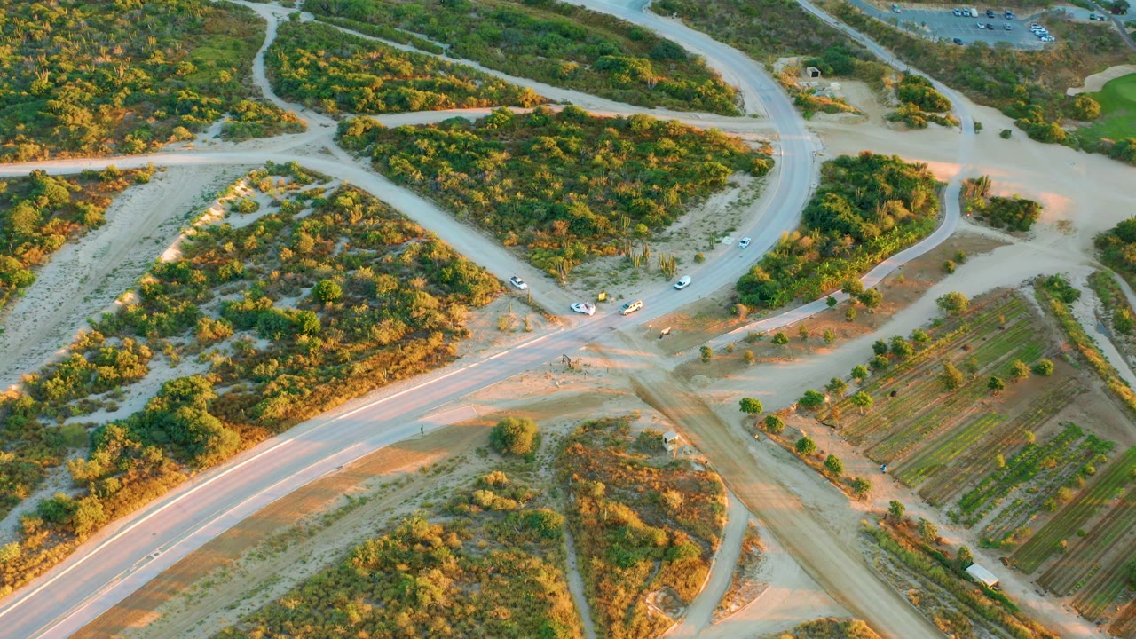 Drone frame in Diamante Cabo San Lucas with cars positioned around a dusty crossing, a curved paved road at lower left, scrub-covered land above, and no visible people.