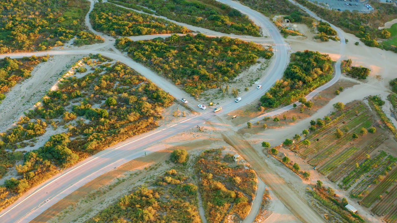High aerial view in Diamante Cabo San Lucas showing the paved road crossing dirt access paths, vehicles spread along the junction, field rows to the right, and no visible people.