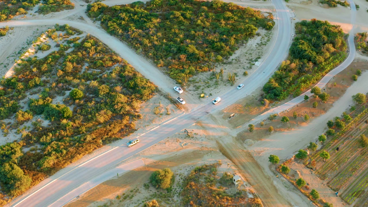 Aerial frame in Diamante Cabo San Lucas with cars spread along the pale roads, a brush-covered mound at left, and no visible people.