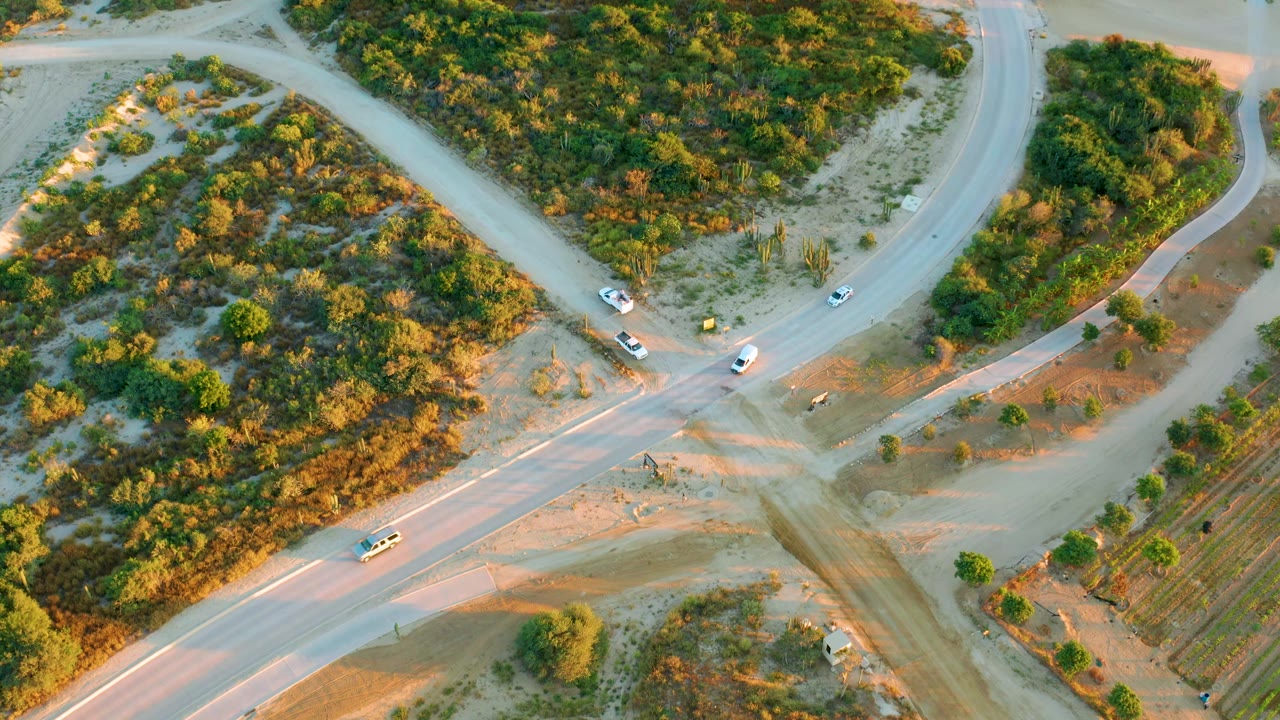 Overhead view in Diamante Cabo San Lucas where vehicles occupy different arms of the dusty junction, with green and brown vegetation filling the surrounding lots and no visible people.