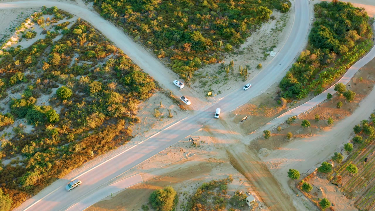Drone frame over Diamante Cabo San Lucas showing a white vehicle near the lower road, other cars near the junction, warm shadows on dirt, and no visible people.