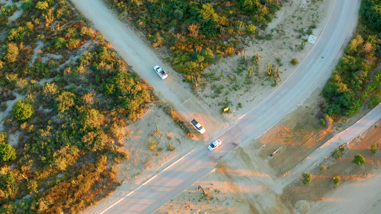 Overhead frame of the Diamante Cabo San Lucas rural roads with cars spaced around the junction, dry sandy areas, dense shrubs, and no visible people.