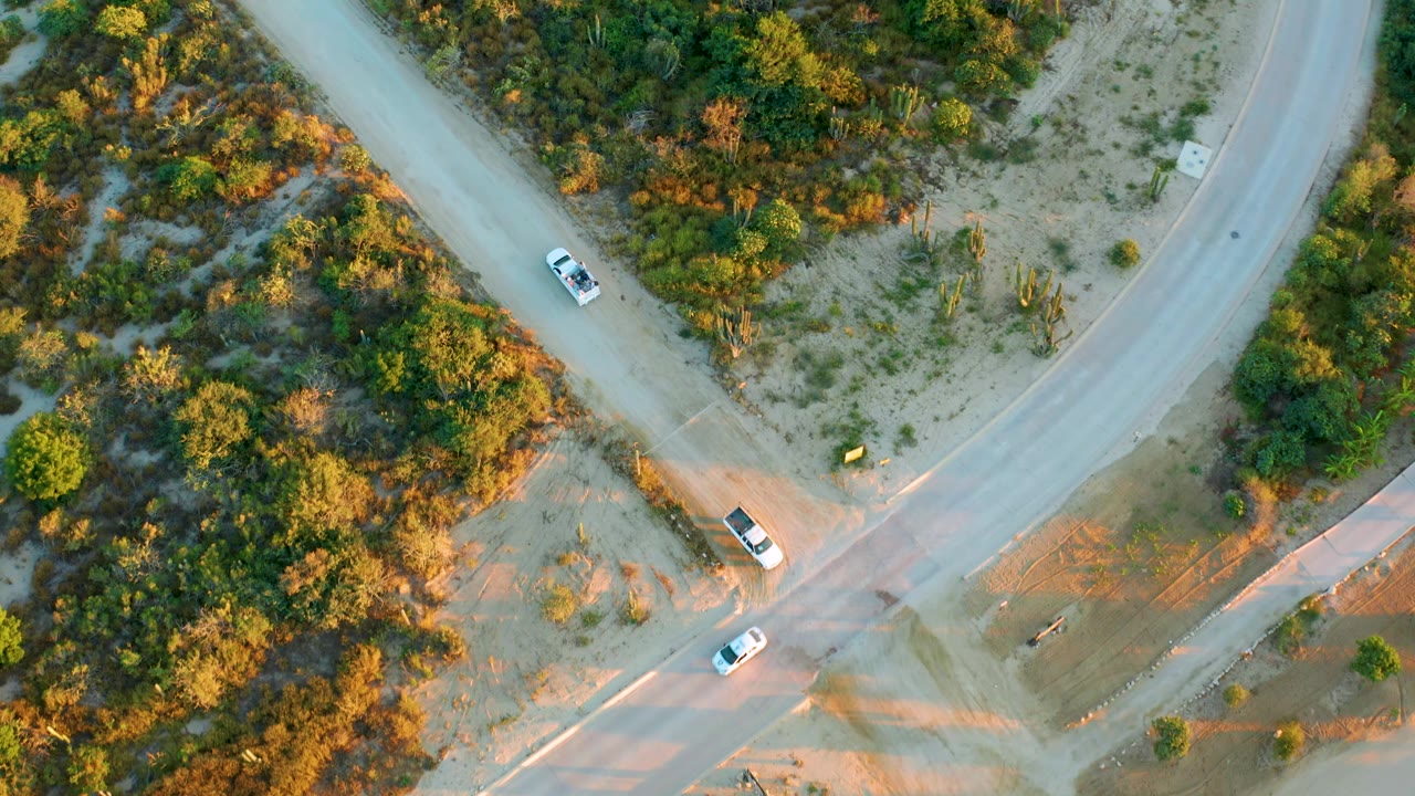 Aerial view in Diamante Cabo San Lucas with vehicles positioned on the intersecting roads, a curved road at the top, scrubland on both sides, and no visible people.
