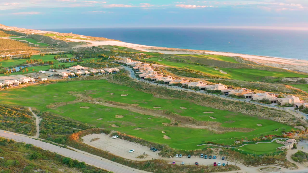 Opening aerial frame over Diamante Cabo San Lucas shows green golf fairways, sand traps, a small parking lot, low buildings, sandy beach, blue ocean, and no visible people.