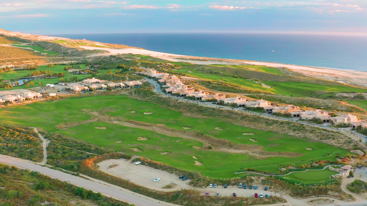 Wide drone view of golf fairways, parking lot, beach, and ocean.