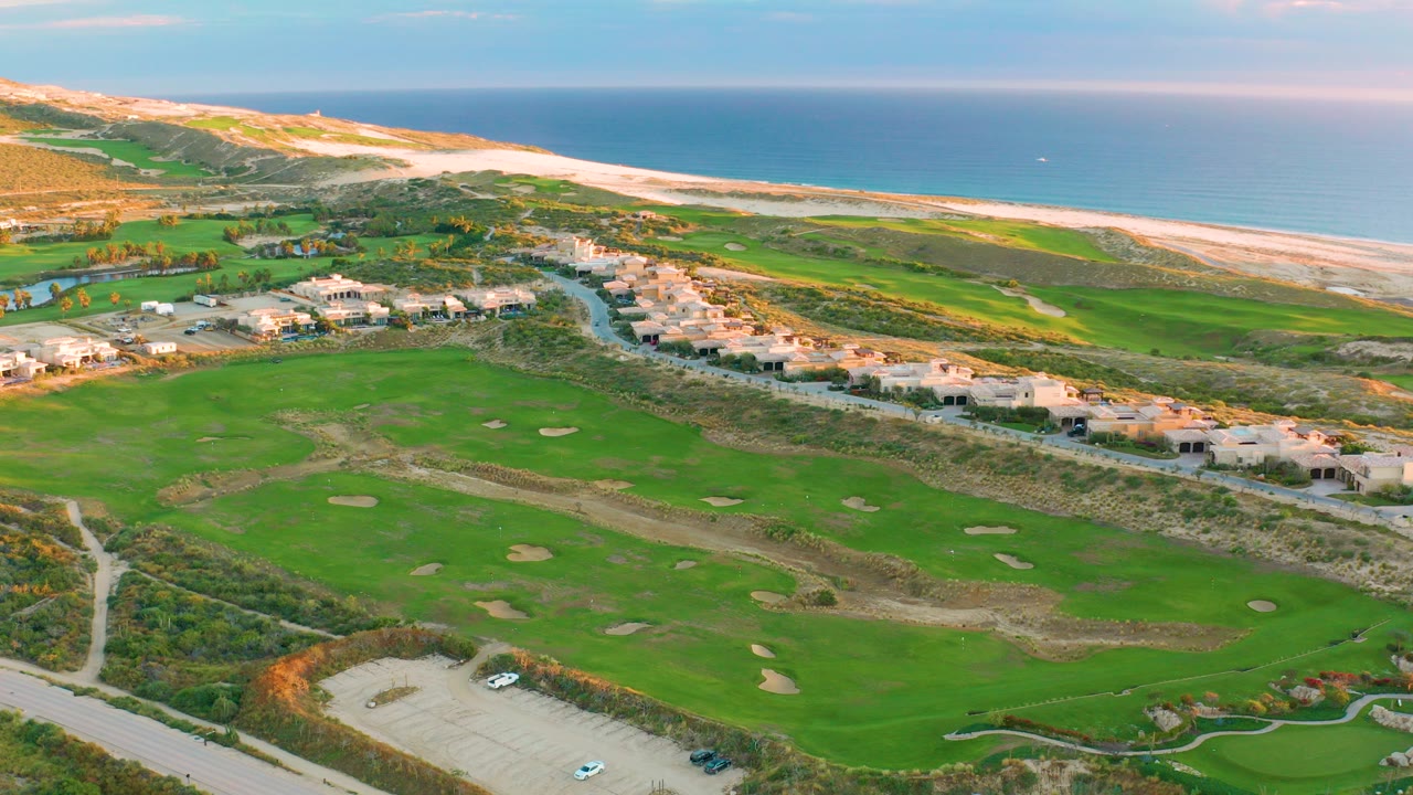 High aerial view over Diamante Cabo San Lucas showing green fairways, sand traps, residential buildings, dunes, beach, blue ocean, and no visible people at the start of the clip.