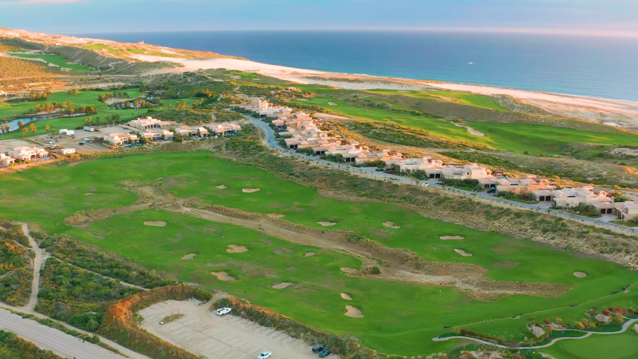 Drone aerial over Diamante Cabo San Lucas showing the course from parking and shrub edges to central fairways, residential roofs, beach dunes, ocean water, and no visible people.