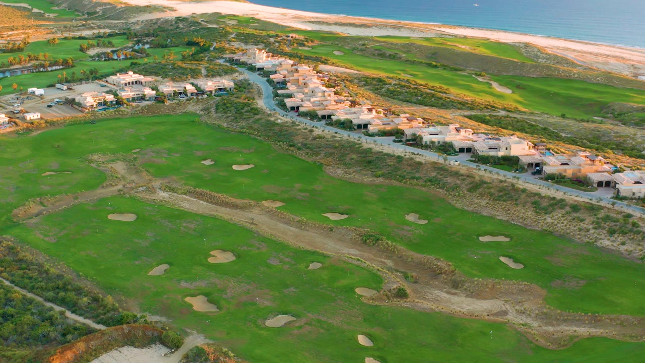 Oblique aerial view over Diamante Cabo San Lucas with green golf fairways and sand traps below, coastal homes along a road, beach and ocean beyond, and no visible people.