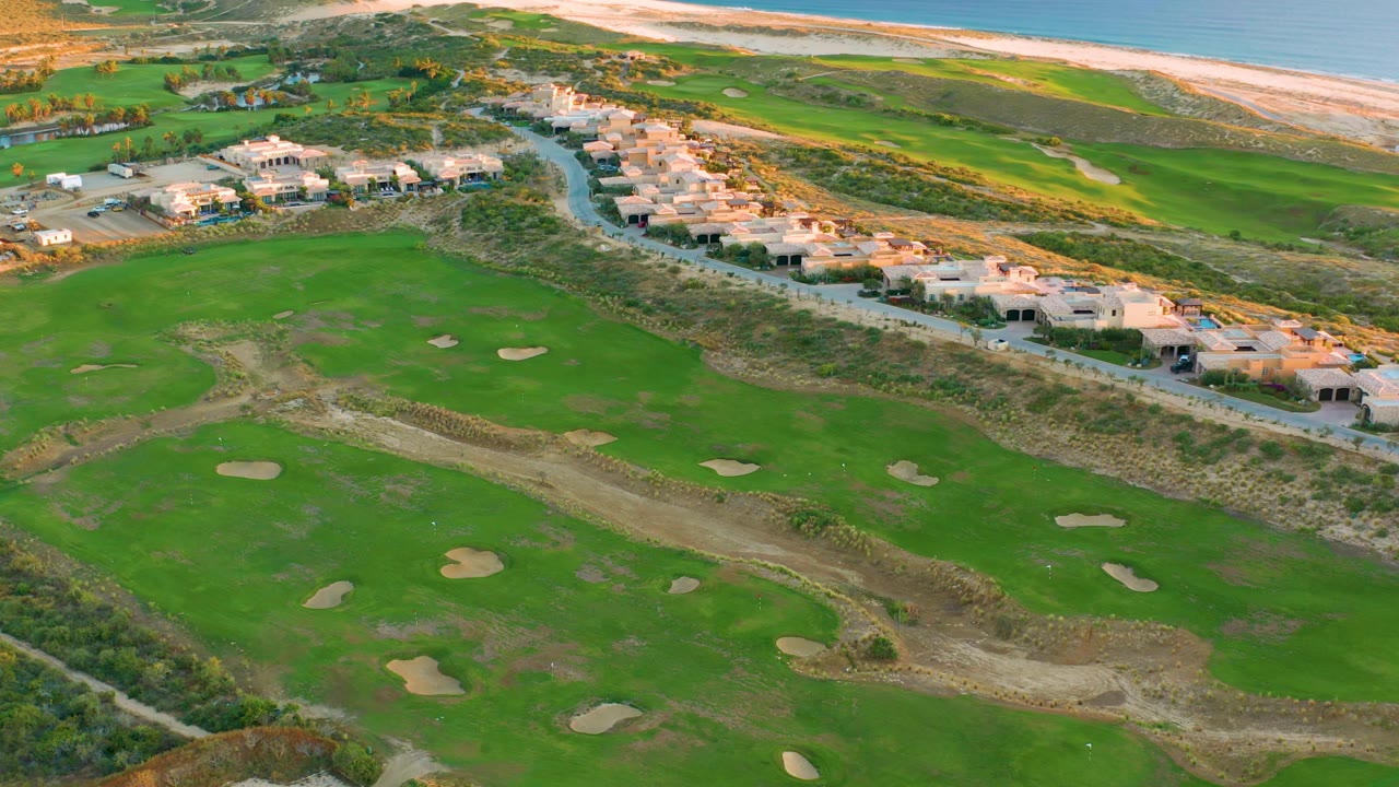 Aerial frame over the Diamante Cabo San Lucas golf course with multiple sand traps across the green, coastal houses beside the road, beach at the top of frame, ocean, and no visible people.