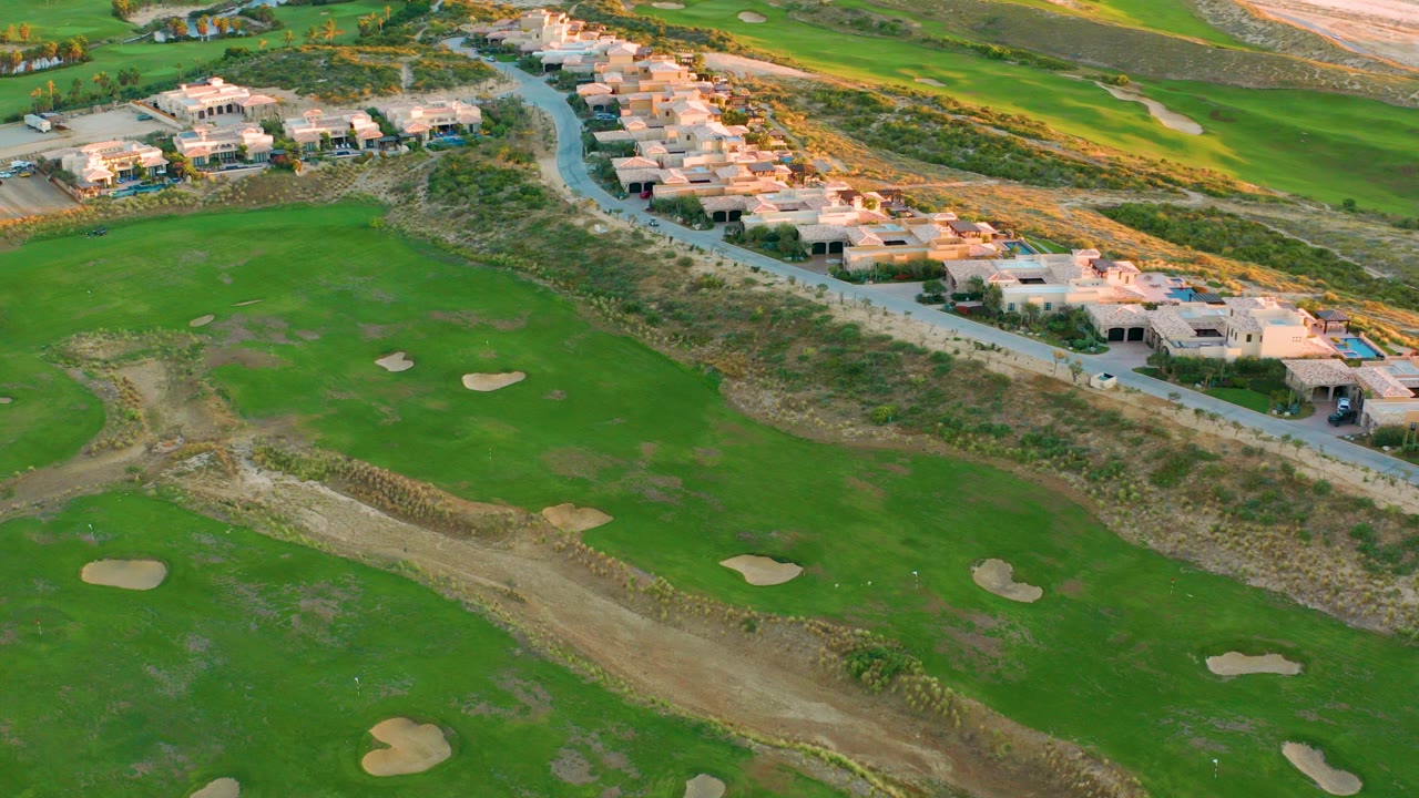 High aerial view over Diamante Cabo San Lucas with green golf fairways, several sand traps, a curving road, light-colored coastal homes, and no visible people.
