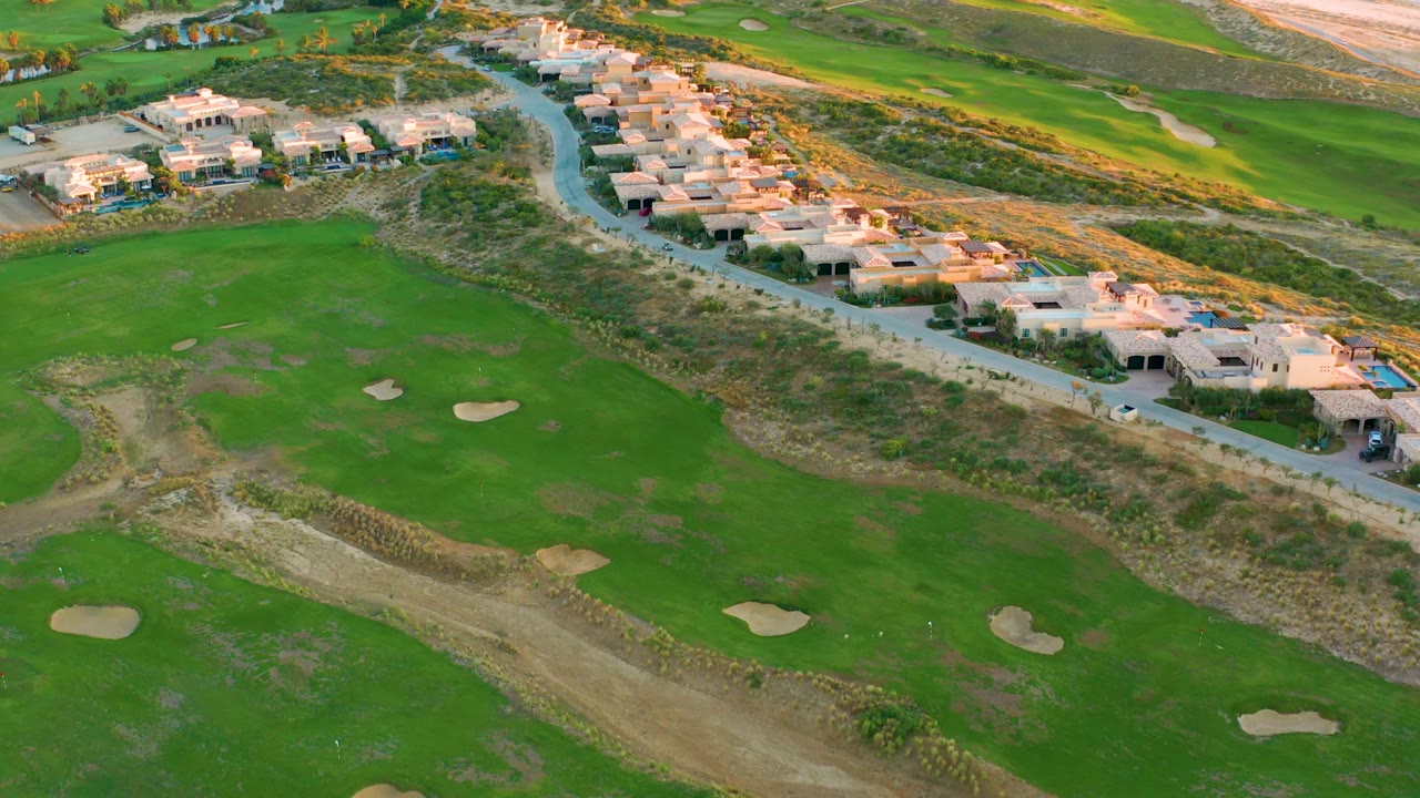 Oblique aerial view in Diamante Cabo San Lucas with fairways and sand traps in the foreground, homes along the road, sandy beach and ocean starting to show at the top, and no visible people.