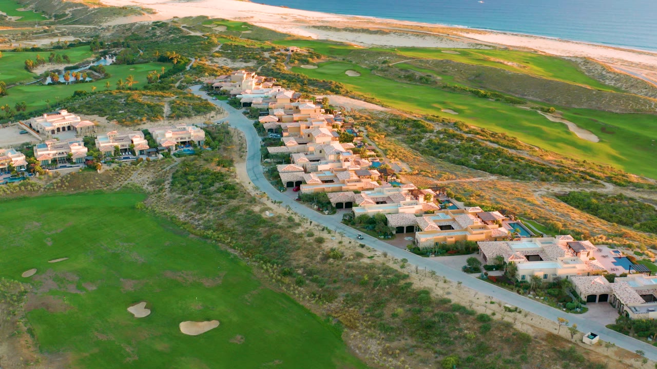 At 0:00, an elevated drone view over Diamante Cabo San Lucas shows villas along a curved road, green golf fairways, sandy dunes, beach, and blue ocean, with no visible people.