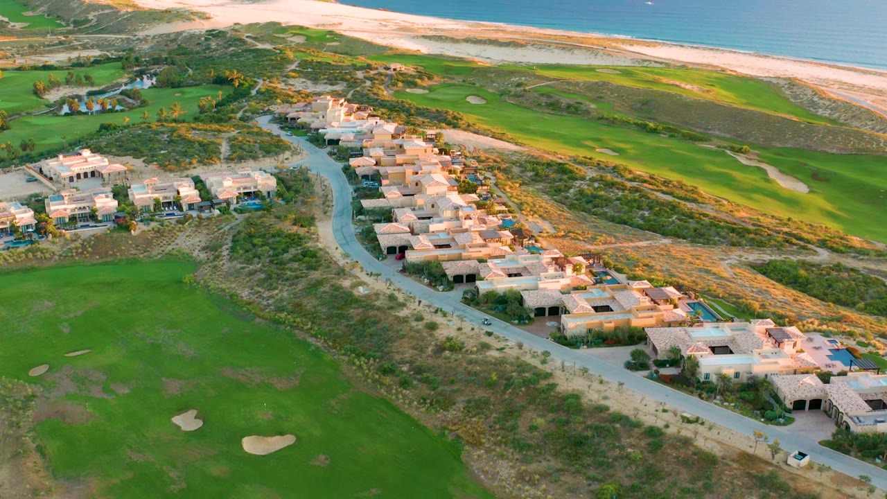 At 0:01, the aerial composition holds over Diamante Cabo San Lucas with the villa row centered, golf greens at left and lower frame, dunes beyond, and a long sandy beach meeting the ocean, with no visible people.