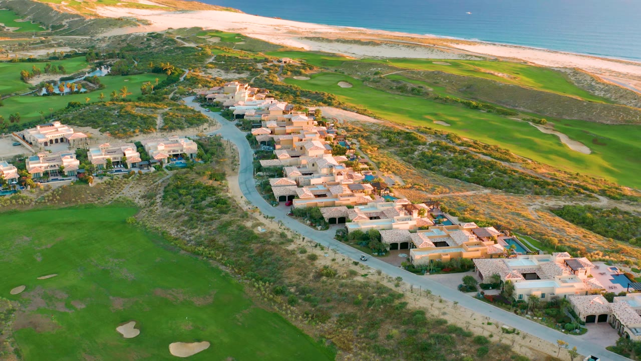 At 0:02, the drone view shows Diamante Cabo San Lucas villas running diagonally through the frame, with golf course grass and bunkers below and the dune-backed beach and ocean stretching across the top, with no visible people.