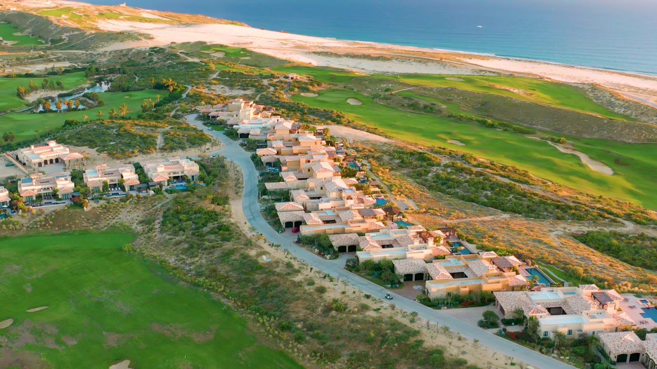 Aerial over curved road, villas, green course, dunes, and beach.