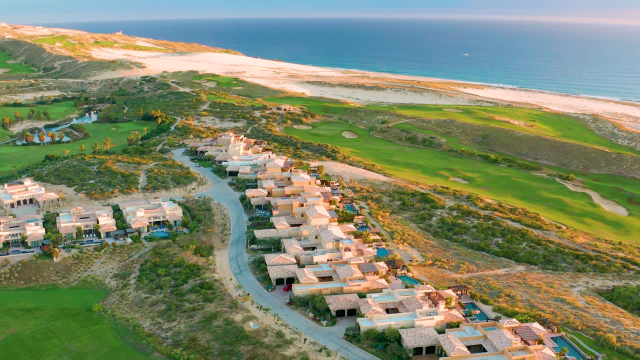Aerial frame over Diamante Cabo San Lucas showing tan residential roofs and a winding road below, green golf fairways and sand traps beyond, pale dunes, sandy beach, calm blue ocean, and no visible people.