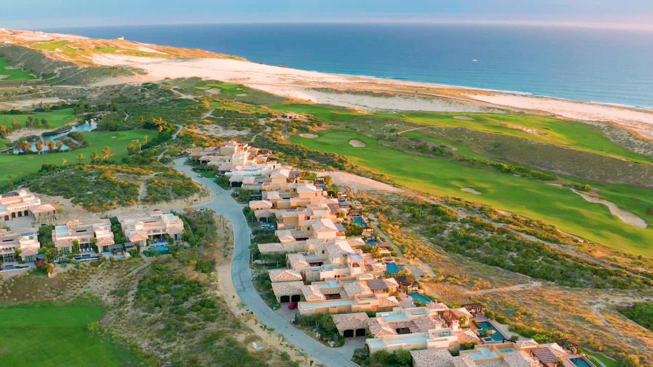 Aerial view in Diamante Cabo San Lucas with the road and clustered houses shifted slightly right, bright fairways and bunkers running across the midground, a long sandy beach and blue ocean beyond, and no visible people.