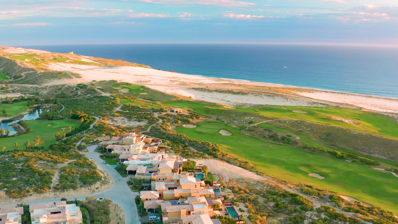 Aerial view in Diamante Cabo San Lucas with houses grouped at the bottom, a curved green fairway and sand traps in the midground, beach and ocean beyond, and no visible people.