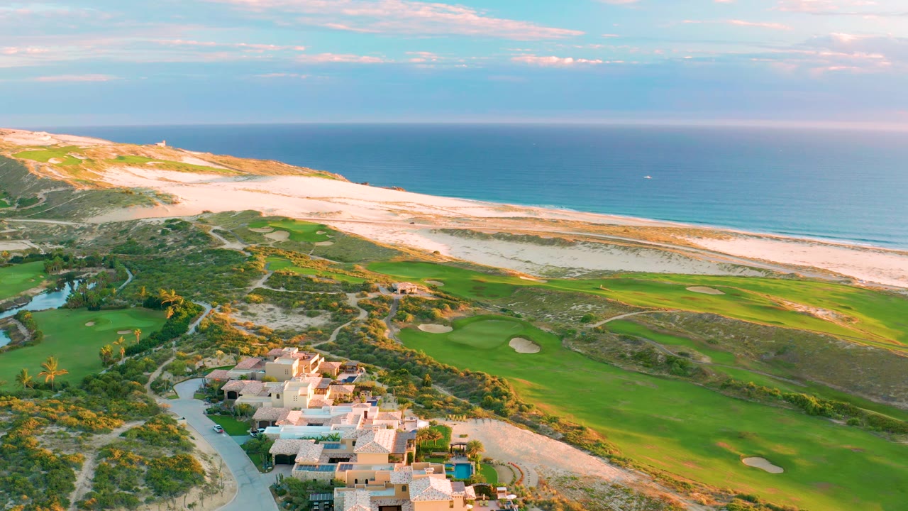 Diamante Cabo San Lucas aerial view with the villa cluster lower in frame, bright fairways and bunkers stretching toward pale dunes, and the ocean horizon across the top, with no visible people.