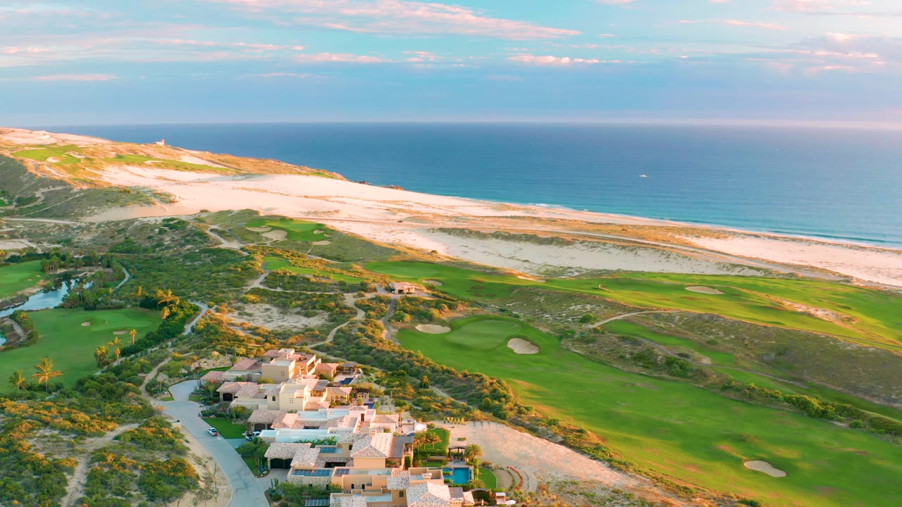 Aerial view at Diamante Cabo San Lucas with resort villas near the lower center, a winding road and green golf course below the dunes, and calm ocean filling the background, with no visible people.