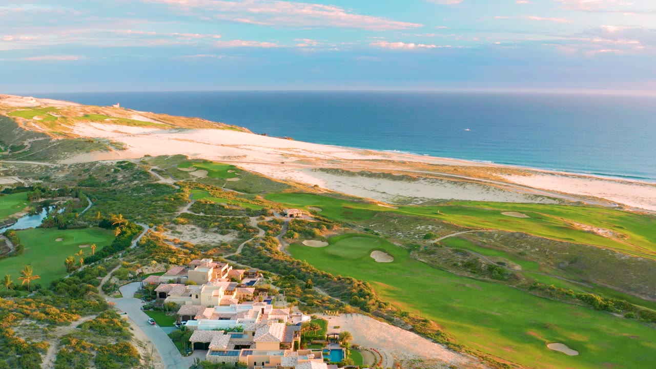 Wide aerial of green golf fairways below beach dunes and ocean
