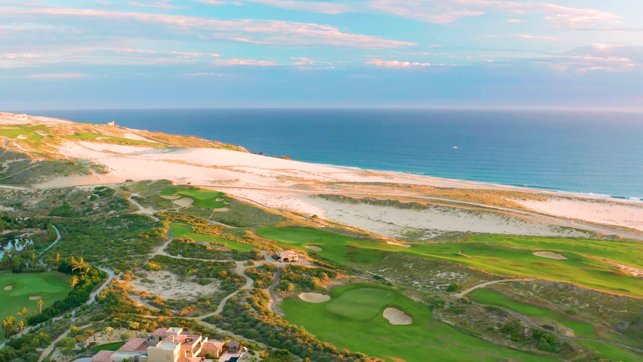 Drone view at Diamante Cabo San Lucas with fairways and bunkers in the lower frame, sandy dunes and beach across the center, ocean beyond, and no visible people.