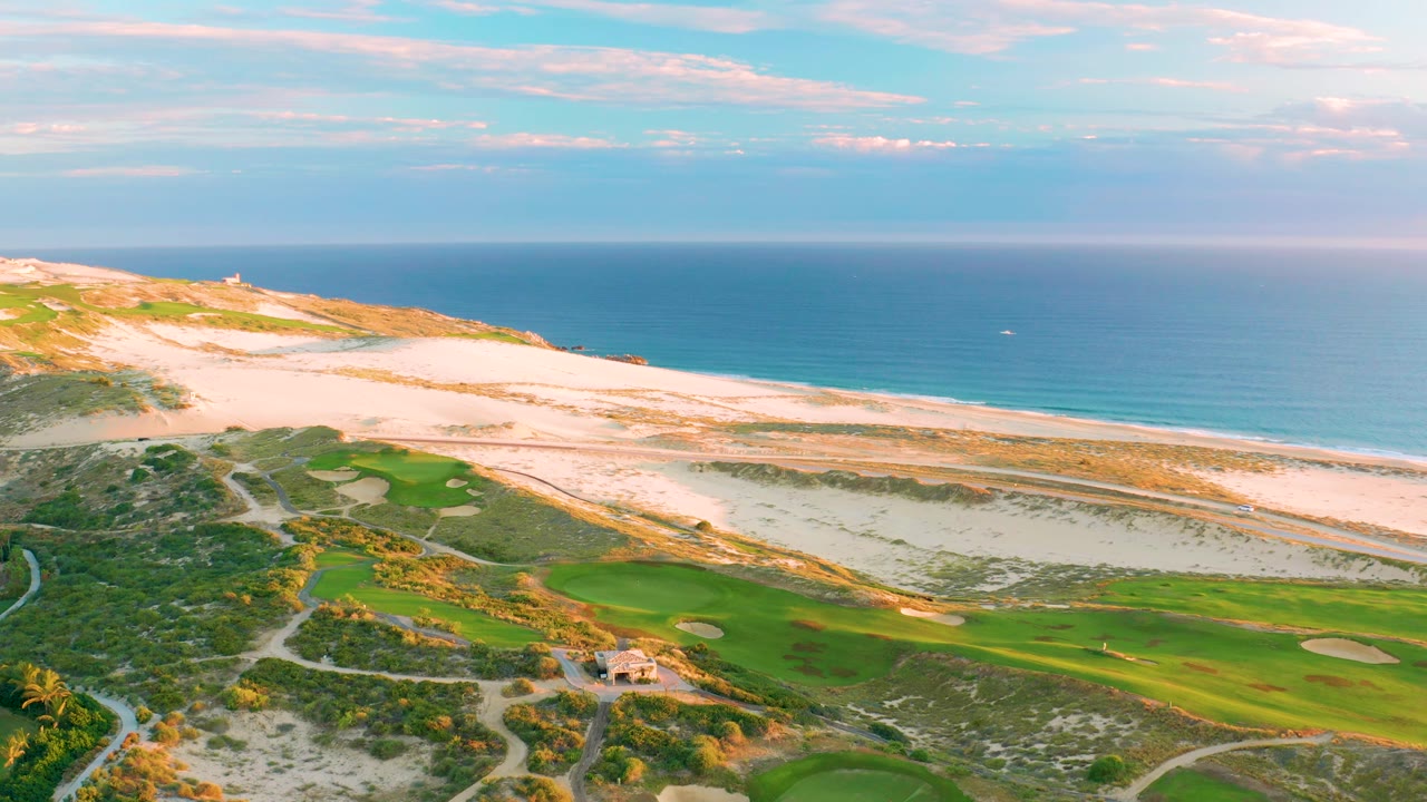 Aerial frame at Diamante Cabo San Lucas with the course and sand traps at the bottom, a long sandy beach across the middle, open blue ocean behind it, and no visible people.