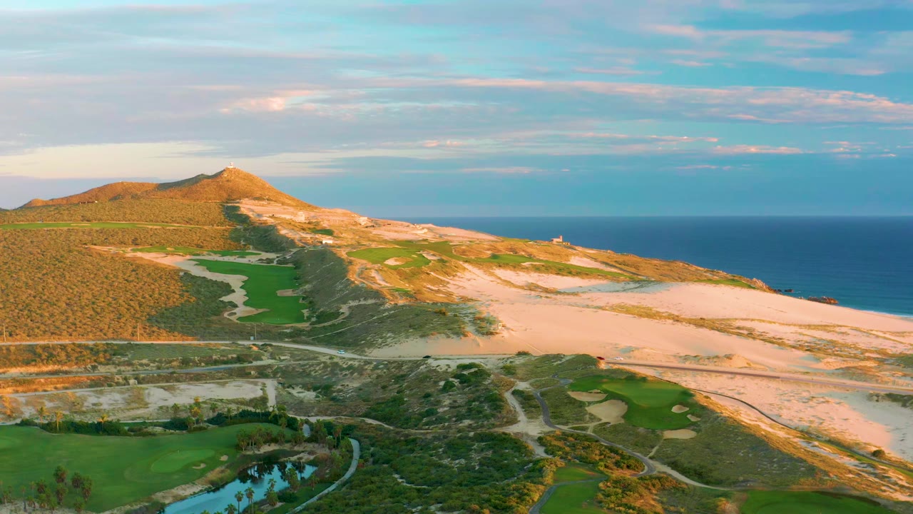 Aerial view of coastal dunes, green fields, a hill, and ocean horizon.