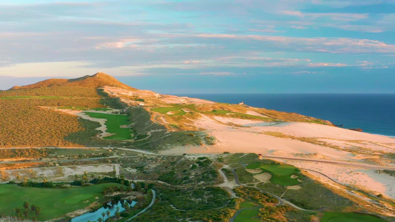 Aerial frame at Diamante Cabo San Lucas with green fairway-like land in the foreground, pale sand dunes across the center, a sunlit hill at left, and no visible people.