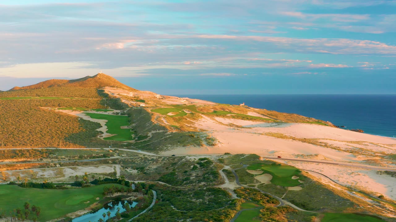 Aerial frame at Diamante Cabo San Lucas showing the sunlit ridge, broad sandy terrain, green fields, a small reflective water feature near the bottom, and no visible people.