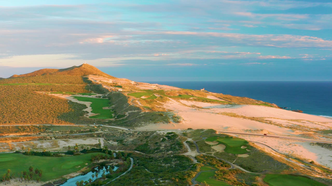 Aerial view of coastal dunes, golf course, ocean, and hill in warm light.