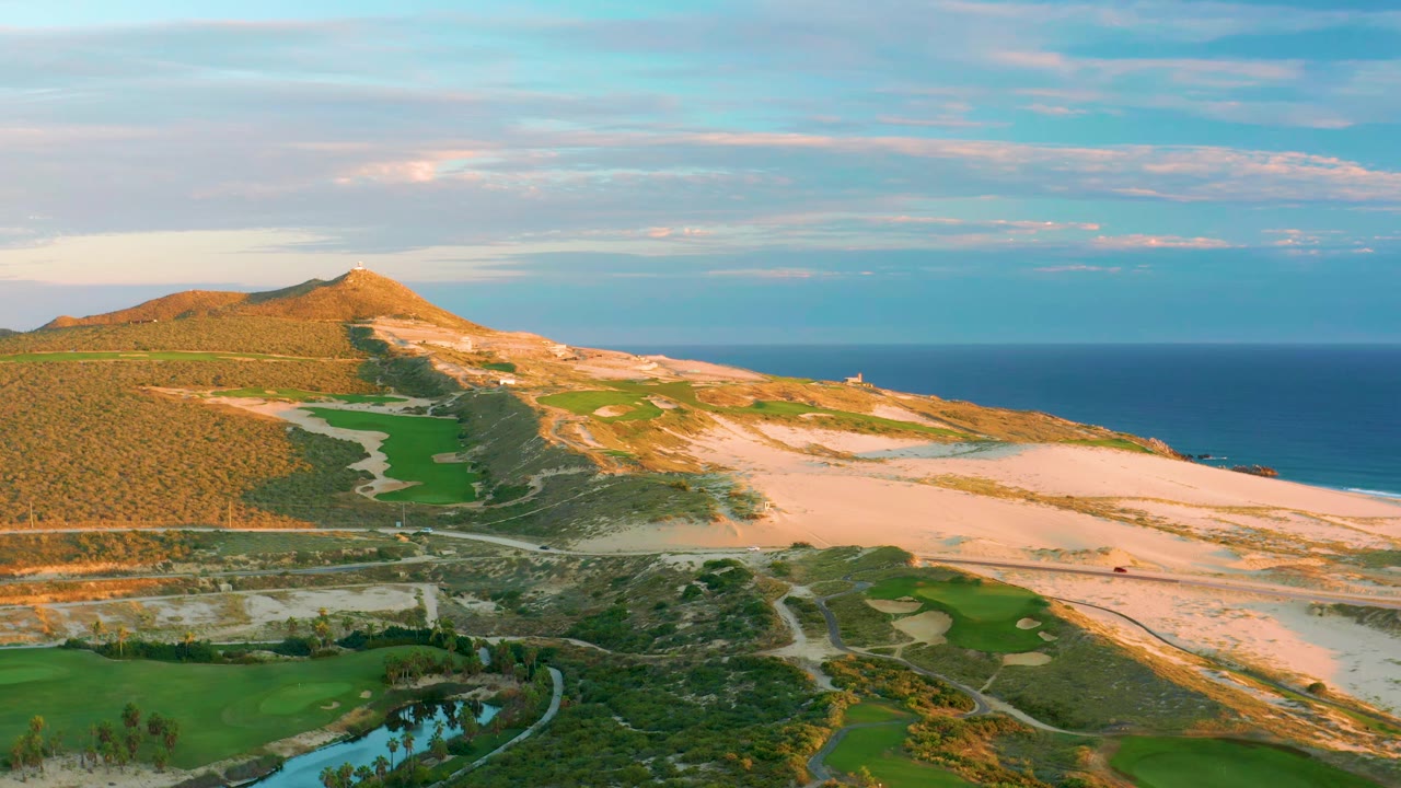 A second aerial view of Diamante Cabo San Lucas keeps the golf course and water channel low in frame while sandy dunes, the hill, and ocean horizon fill the distance, with no visible people.