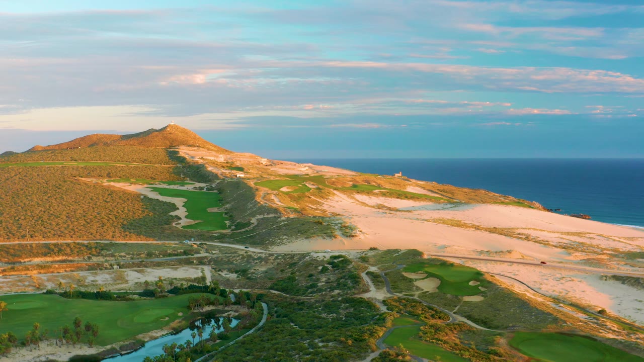 Mid-clip aerial frame over Diamante Cabo San Lucas showing green fairways at the lower left, a pale dune slope with tracks across the center, the hill in warm light, and the ocean to the right, with no visible people.