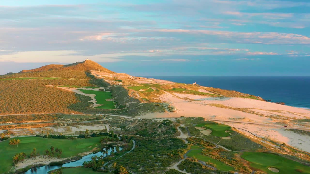 Aerial view at Diamante Cabo San Lucas with fairways and cart paths in the lower frame, pale dunes across the middle, and blue ocean stretching across the background with no visible people.