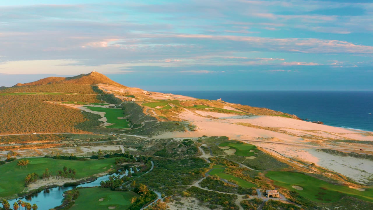Aerial frame at Diamante Cabo San Lucas showing sandy dunes, green fields, a pond in the lower left, a low hill, ocean horizon, pastel clouds, and no visible people.